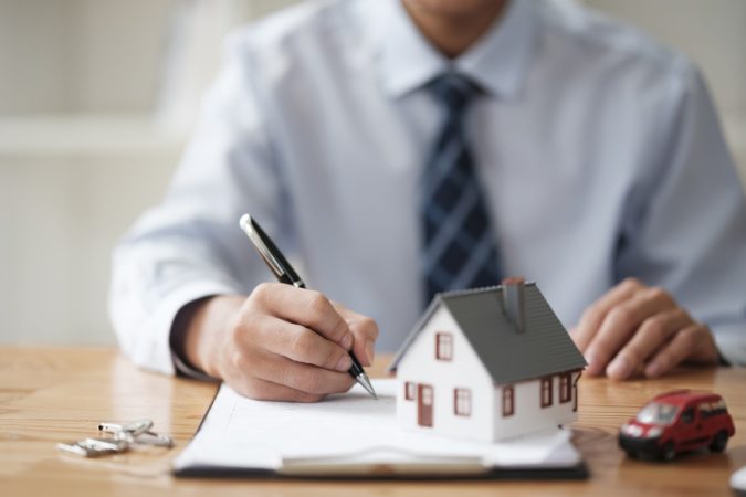 A businessman signing a real estate contract with house and car models on the table, representing property and vehicle purchase agreements.