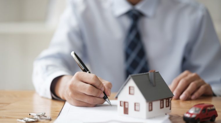 A businessman signing a real estate contract with house and car models on the table, representing property and vehicle purchase agreements.